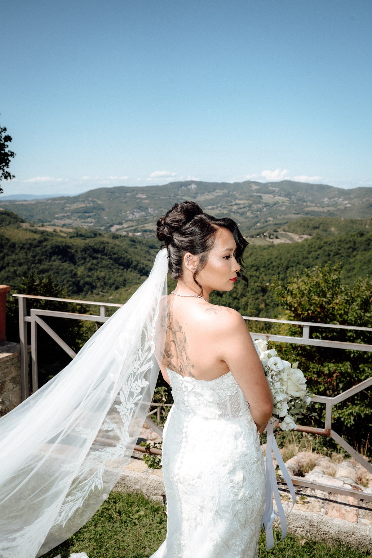 Braut mit langem Schleier blickt über die Schulter vor Hügellandschaft, Hochzeitsfoto in Italien